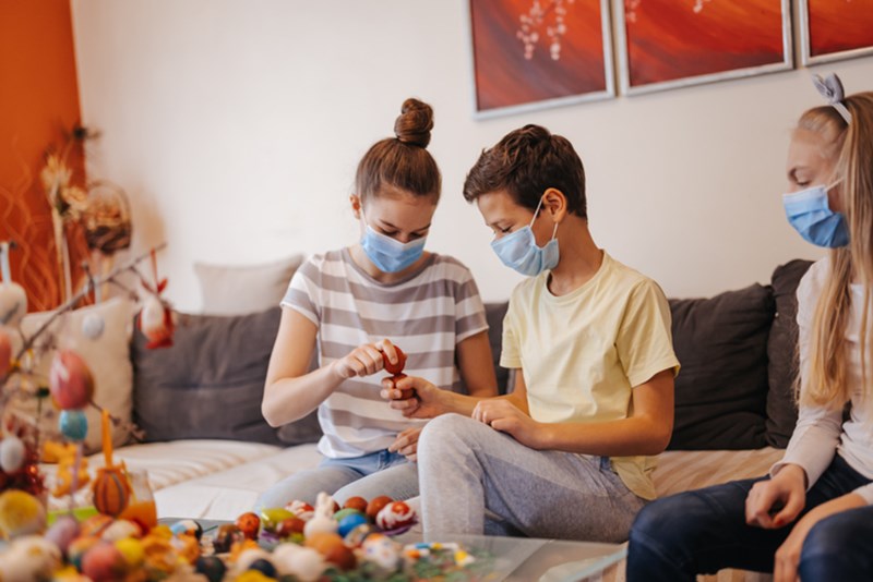 THREE CHILDREN WITH PROTECTIVE FACE MASKS BREAKING EASTER EGGS TOGETHER